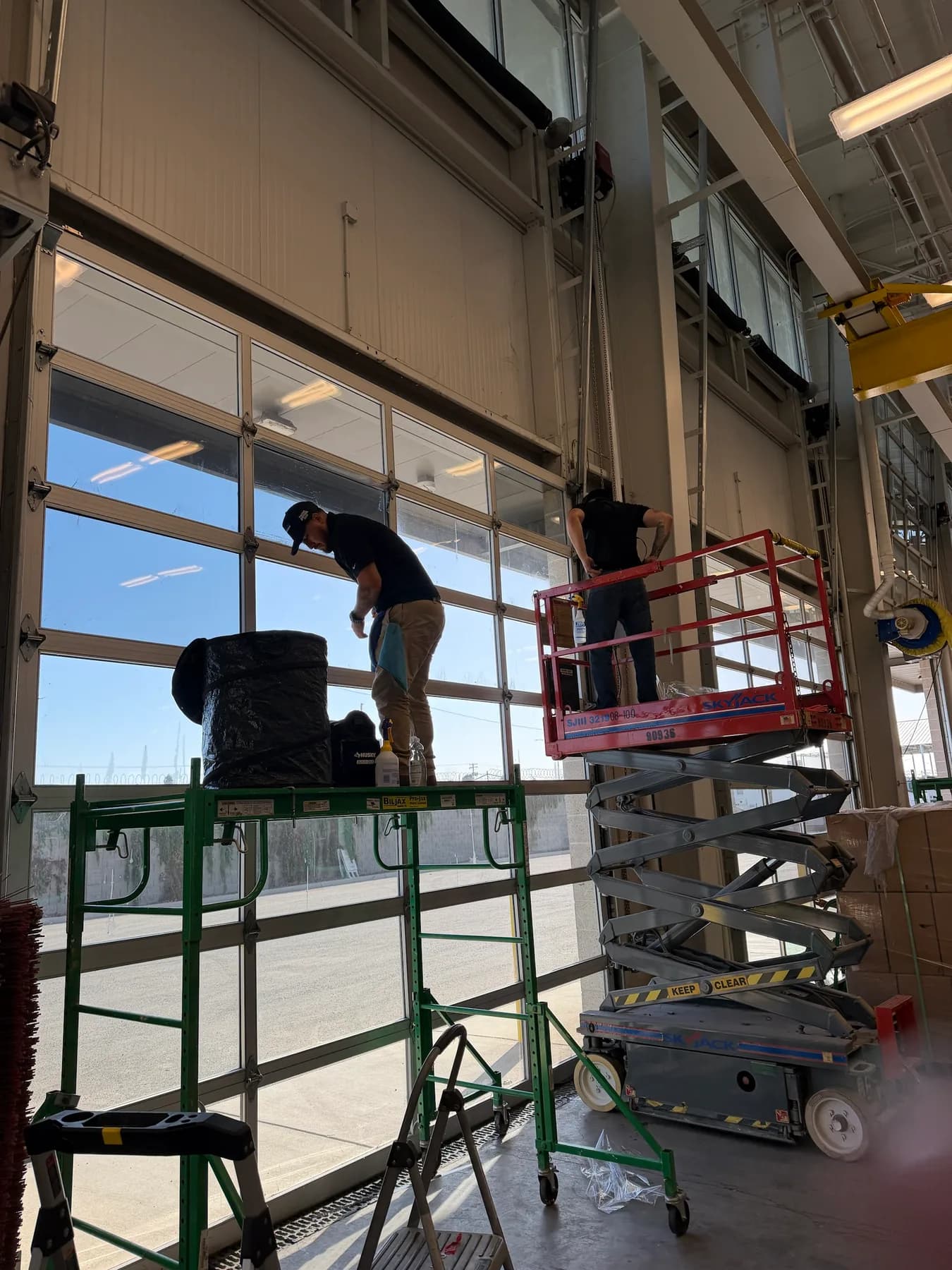 Window film installers on a scissor lift and rolling scaffold filming the upper panes of a glass bay door